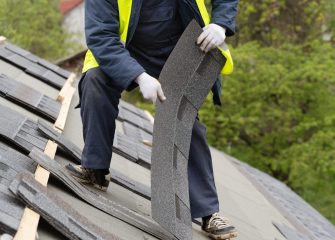 Professional and skilled roofer worker in special protective work wear installing asphalt or bitumen shingle on top of the new roof under construction residential building